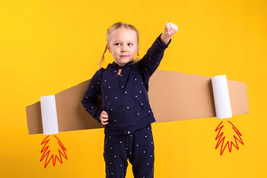 A little child girl is wearing homemade cardboard flying wings, pretending to be a pilot for a craft, imagination or exploration concept. Studio photography on a yellow background
