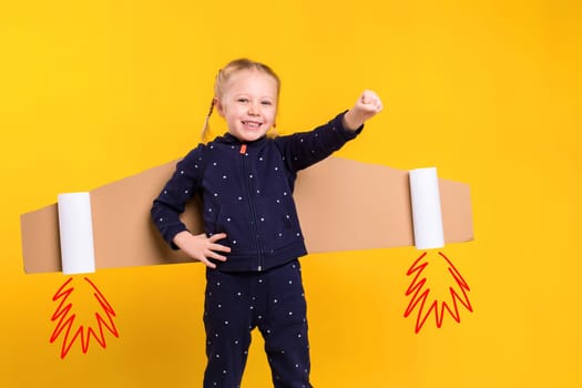 A little child girl is wearing homemade cardboard flying wings, pretending to be a pilot for a craft, imagination or exploration concept. Studio photography on a yellow background