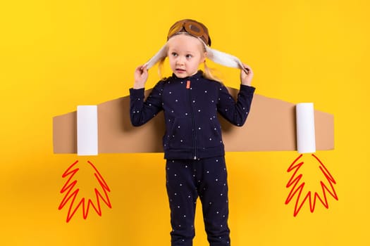 A little child girl is wearing homemade cardboard flying wings, pretending to be a pilot for a craft, imagination or exploration concept. Studio photography on a yellow background