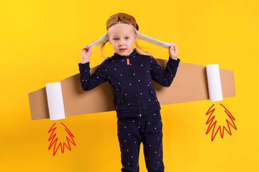 A little child girl is wearing homemade cardboard flying wings, pretending to be a pilot for a craft, imagination or exploration concept. Studio photography on a yellow background