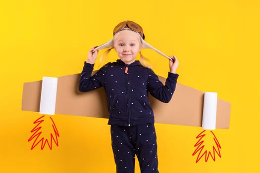 Freedom, girl playing to be airplane pilot, funny little girl with aviator cap and glasses, carries wings made of brown cardboard as an airplane. Studio photography on a yellow background