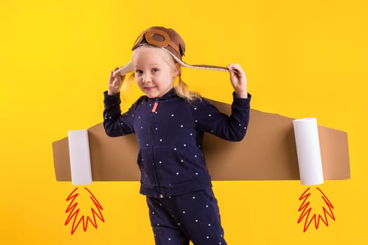 Freedom, girl playing to be airplane pilot, funny little girl with aviator cap and glasses, carries wings made of brown cardboard as an airplane. Studio photography on a yellow background