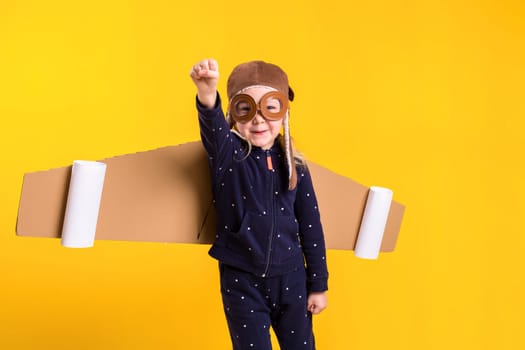 Freedom, girl playing to be airplane pilot, funny little girl with aviator cap and glasses, carries wings made of brown cardboard as an airplane. Studio photography on a yellow background