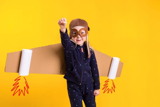 Freedom, girl playing to be airplane pilot, funny little girl with aviator cap and glasses, carries wings made of brown cardboard as an airplane. Studio photography on a yellow background