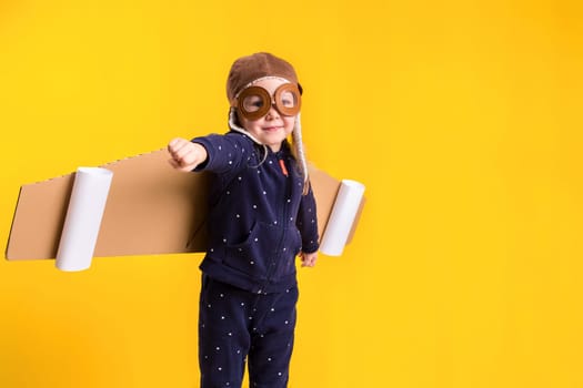 Freedom, girl playing to be airplane pilot, funny little girl with aviator cap and glasses, carries wings made of brown cardboard as an airplane. Studio photography on a yellow background