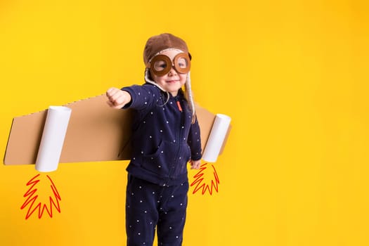 Freedom, girl playing to be airplane pilot, funny little girl with aviator cap and glasses, carries wings made of brown cardboard as an airplane. Studio photography on a yellow background