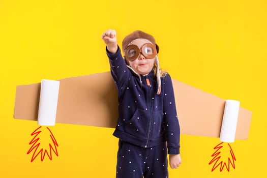 Freedom, girl playing to be airplane pilot, funny little girl with aviator cap and glasses, carries wings made of brown cardboard as an airplane. Studio photography on a yellow background