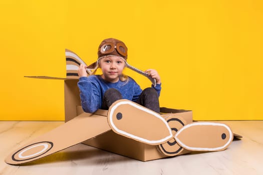 Little dreamer girl playing with a cardboard airplane. Childhood. Fantasy, imagination. Studio photography on a yellow background. Imagination or exploration concept