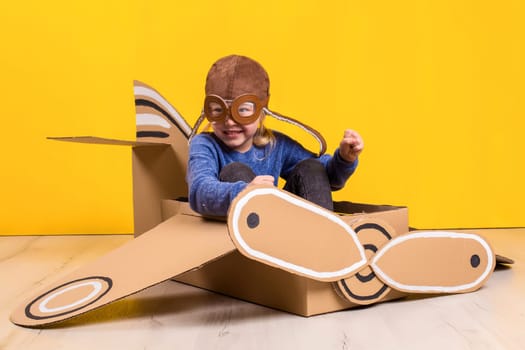Little dreamer girl playing with a cardboard airplane. Childhood. Fantasy, imagination. Studio photography on a yellow background. Imagination or exploration concept