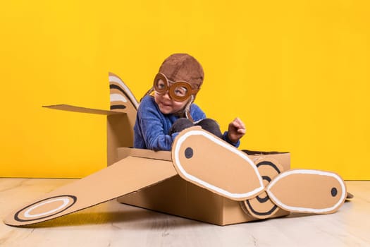 Little dreamer girl playing with a cardboard airplane. Childhood. Fantasy, imagination. Studio photography on a yellow background. Imagination or exploration concept