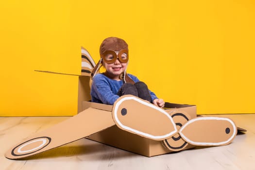 Little dreamer girl playing with a cardboard airplane. Childhood. Fantasy, imagination. Studio photography on a yellow background. Imagination or exploration concept