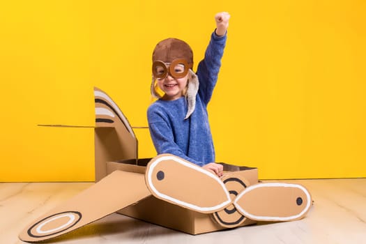 Little dreamer girl playing with a cardboard airplane. Childhood. Fantasy, imagination. Studio photography on a yellow background. Imagination or exploration concept