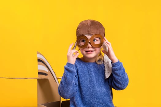 Little dreamer girl playing with a cardboard airplane. Childhood. Fantasy, imagination. Studio photography on a yellow background. Imagination or exploration concept