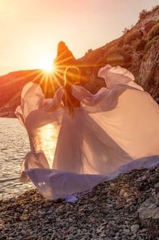 Mysterious woman silhouette long hair walks on the beach ocean water, sea nymph wind listens to the wave. Throws up a long white dress, a divine sunset. Artistic photo from the back without a face.