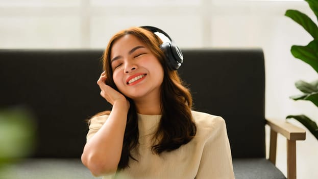 Relaxed millennial woman enjoying favorite track, listening to music through headphones while sitting in cozy living room.