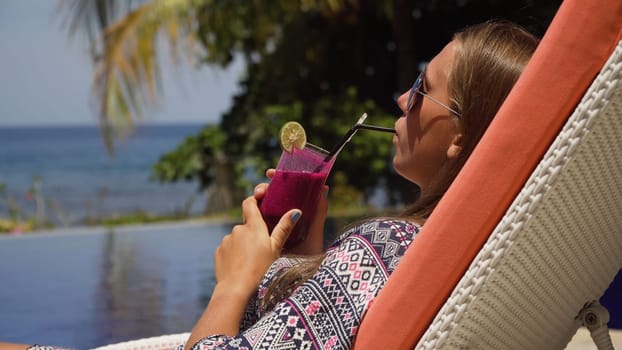 Beautiful young girl with glass of cocktail relaxing near swimming pool. Girl in sunglasses with cocktail in seafront near the bassin amongst the palm trees on the background of the sea. Travel, vacation concept.