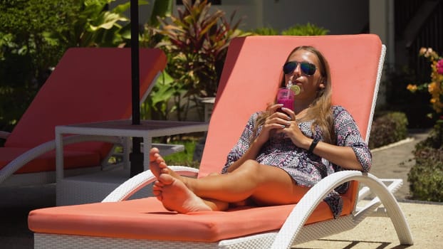 Beautiful young girl with glass of cocktail relaxing near swimming pool. Girl in sunglasses with cocktail in seafront near the bassin amongst the palm trees on the background of the sea. Travel, vacation concept.