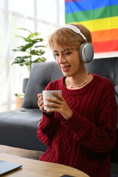 Pretty young gay man wearing headphone drinking coffee watching videos, browsing internet on laptop at home.