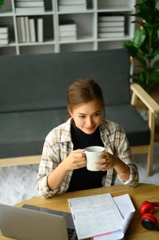 Young female freelancer sitting on floor in living room with coffee cup and using laptop computer.