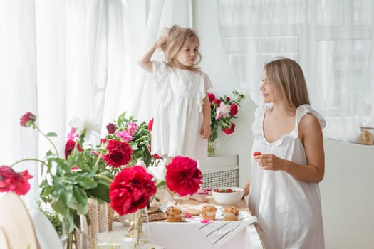 A little blonde girl with her mom on a kitchen countertop decorated with peonies. The concept of the relationship between mother and daughter. Spring atmosphere