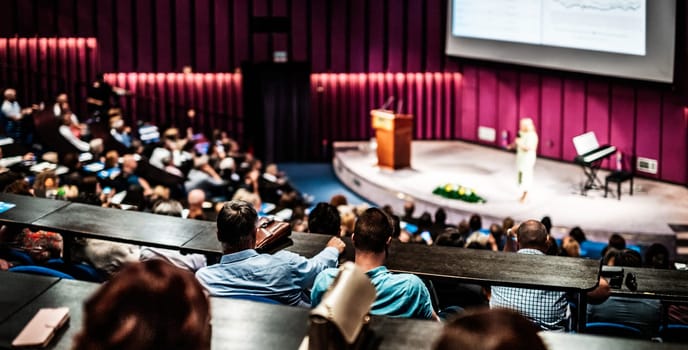 Business and entrepreneurship symposium. Female speaker giving a talk at business meeting. Audience in conference hall. Rear view of unrecognized participant in audience.