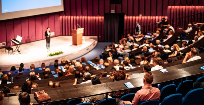 Business and entrepreneurship symposium. Female speaker giving a talk at business meeting. Audience in conference hall. Rear view of unrecognized participant in audience.