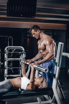 Personal trainer helping a young woman lift a barbell while working out in a gym. Personal trainer with a gorgeous body without a T-shirt in the gym