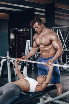 Personal trainer helping a young woman lift a barbell while working out in a gym. Personal trainer with a gorgeous body without a T-shirt in the gym
