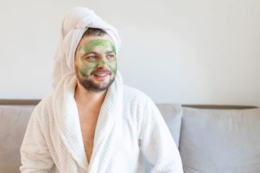 Surprised guy spends hours treating and caring for his skin, applies green face mask to pamper himself, wearing a soft bathrobe and a towel on his head.