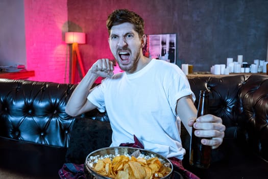 Young fan man watching football game on television celebrating goal happy on sofa couch at home with beer bottle in crazy enthusiastic face expression