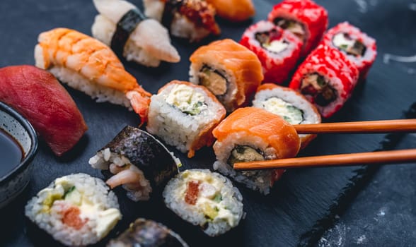Colorful sushi set on black desk with California roll. Person take maki with chopstick. Oriental japanese healthy cuisine with raw fish and rice