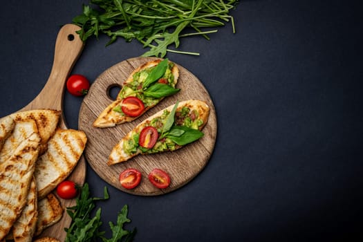 Bruschettas with toast, guacamole, basil and cherry tomatoes on wooden board served with arugula. Mexican snack with french slice bread with avocado, herbs and vegetables on background with copy space