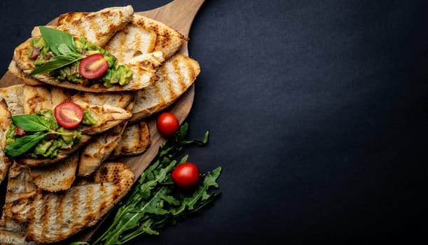 Bruschettas with toast, guacamole, basil and cherry tomatoes on wooden board served with arugula. Beautiful food composition with slice bread, avocado, herbs and vegetables with copyspace background
