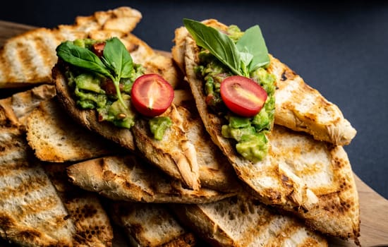Bruschettas french toasts with guacamole, basil and cherry tomatoes on wooden board. Mexican snack antipasti with slice bread, mashed avocado, herbs and vegetables