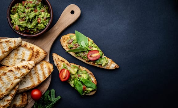 Bruschettas with toast, guacamole, basil and cherry tomatoes on wooden board served with arugula. Beautiful food composition with slice bread, avocado, herbs and vegetables with copyspace background