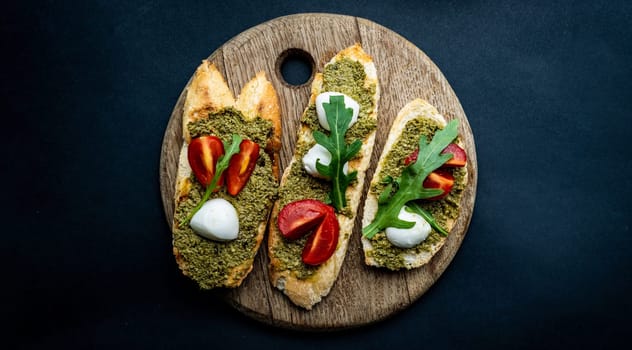 Bruschettas with pesto, tomatoes, mozzarella cheese and basil on wooden board. Italian tosted bread with vegetables and traditional sauce