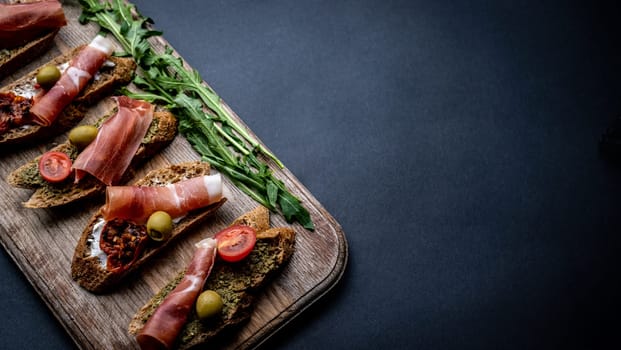 Bruschettas with jamon, olives, pesto, grilled cherry tomatoes served on wooden board with arugula on background with copy space. Mediterranean toasted bread with meat, cheese and vegetables