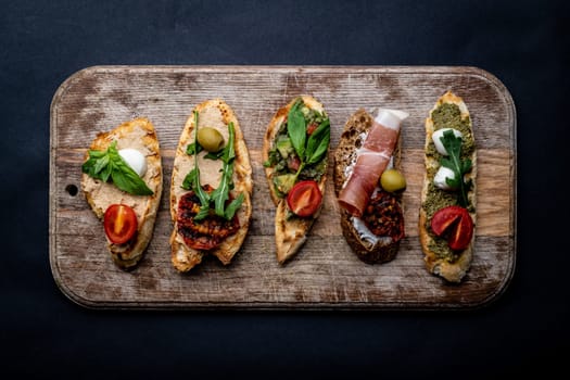 Set of bruschettas with jamon, olives, pesto, grilled and cherry tomatoes, basil served on wooden board with arugula. Traditional mediterranean toasted bread with cheese, meat and vegetables