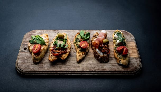 Set of bruschettas with jamon, olives, pesto, grilled and cherry tomatoes, basil served on wooden board with arugula. Traditional mediterranean toasted bread with cheese, meat and vegetables