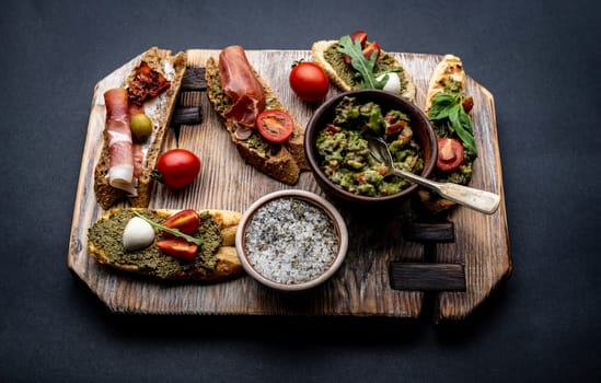 Set of bruschettas with jamon, olives, pesto, tomatoes and basil served on wooden board with guacamole and salt with herbs. Stylish food composition with mediterranean antipasti
