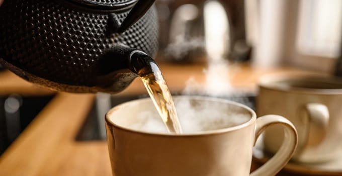 Pouring hot black tea from metal teapot at the kitchen. Steam comes out of the cup. T