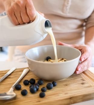 Girl puts milk to oatmeal with blueberry in bowl plate for delicious cereal breakfast at kitchen. Woman preparing granola with berry and latte for morning healthy diet meal