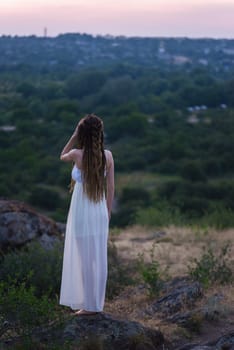 A very beautiful girl in a white dress with dreadlocks is standing on a rock with her arms outstretched against a cloudy sky at sunset light.