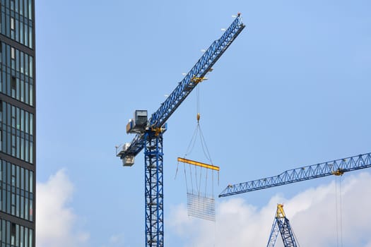 Large tall industrial crane at skyscraper building site, steel bars grid hanging under when moved, clear sky background.