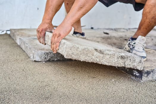 Installing new pavement or floor outside from large concrete tiles, closeup detail on male worker fitting stone block over sand and gravel base layer.