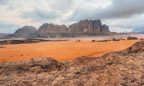 Rocky desert scenery in Wadi Rum desert during overcast morning, camp tents visible in distance.