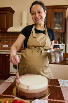 Happy pregnant woman in beige chef's apron, smiling looking at camera, pouring ruby pink chocolate icing over triple chocolate layered mousse dessert with whipped cream and melted Belgian chocolate
