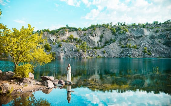 A very beautiful girl in a white dress with dreadlocks is standing on a rock at the lake, at sunrise.