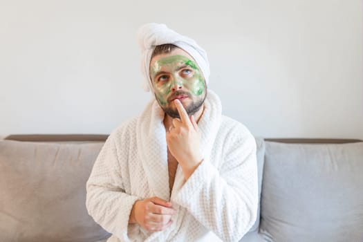 Funny bearded man is having fun with a cosmetic mask on his face made of green clay. Male concept of skincare, humor and spa at home.