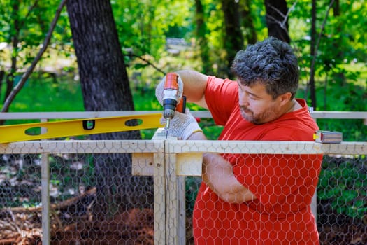 On farm worker uses screwdriver to install metal grid in wooden chicken coop that has been constructed from wood